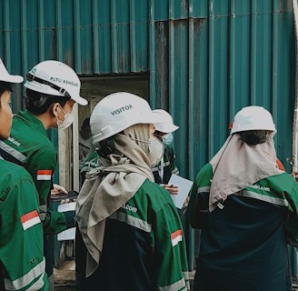 A group of individuals wearing white helmets and green uniform jackets stands near a corrugated metal wall. They appear to be engaged in some activity, possibly an inspection or discussion, holding documents and writing materials. One individual is wearing a headscarf under the helmet.