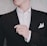 A person is adjusting their black suit jacket, which is worn over a white dress shirt. The hand is lightly touching the jacket, and a white pocket square is neatly tucked into the chest pocket. The background is a neutral, textured wall.